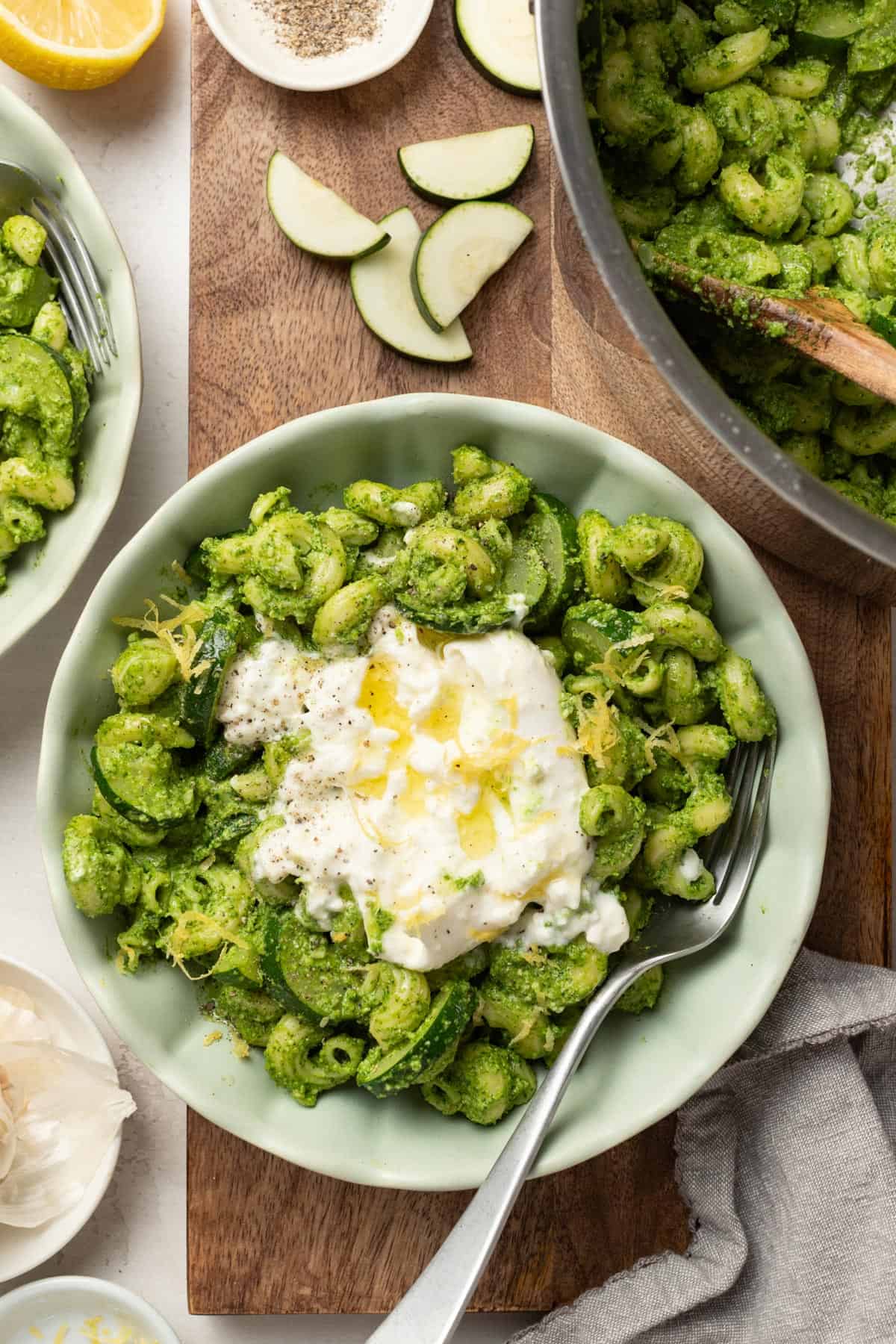 A bowl with burrata pasta and pesto next to a cutting board with spinach, zucchini, and garlic.