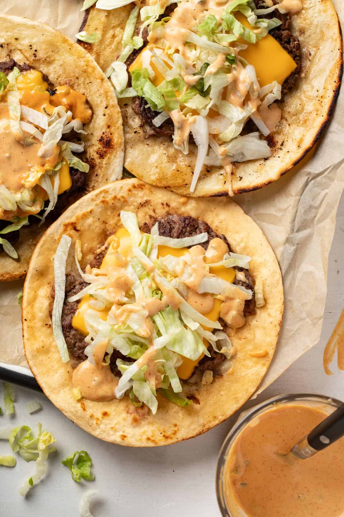 Three burger tacos on a piece of parchment paper, topped with lettuce and burger sauce.