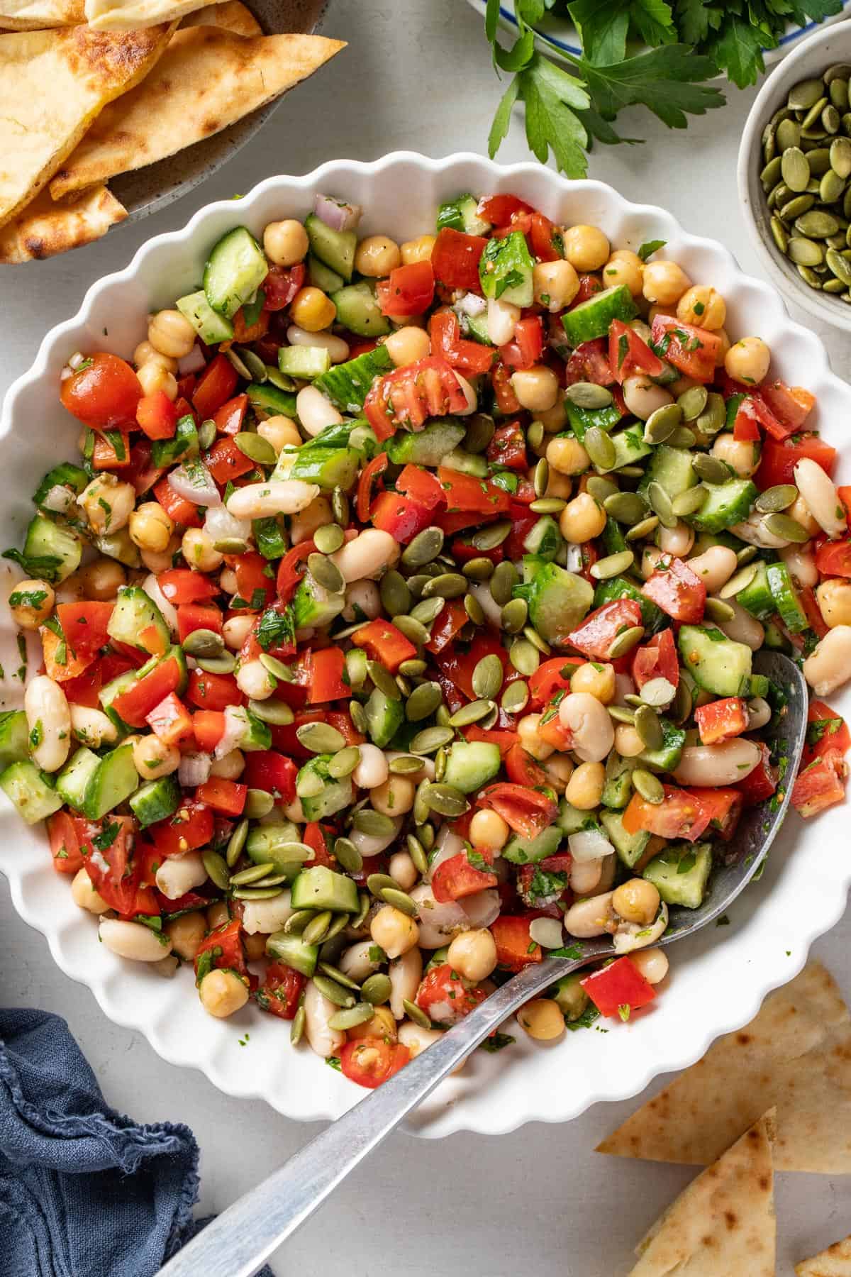 A bean salad with chickpeas, tomatoes, red bell pepper, cucumbers, and cannellini beans in a white bowl.