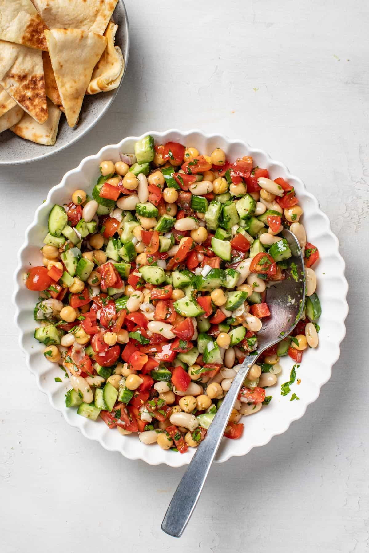 A bean salad mixed together with a spoon next to a bowl of pita bread quarters.