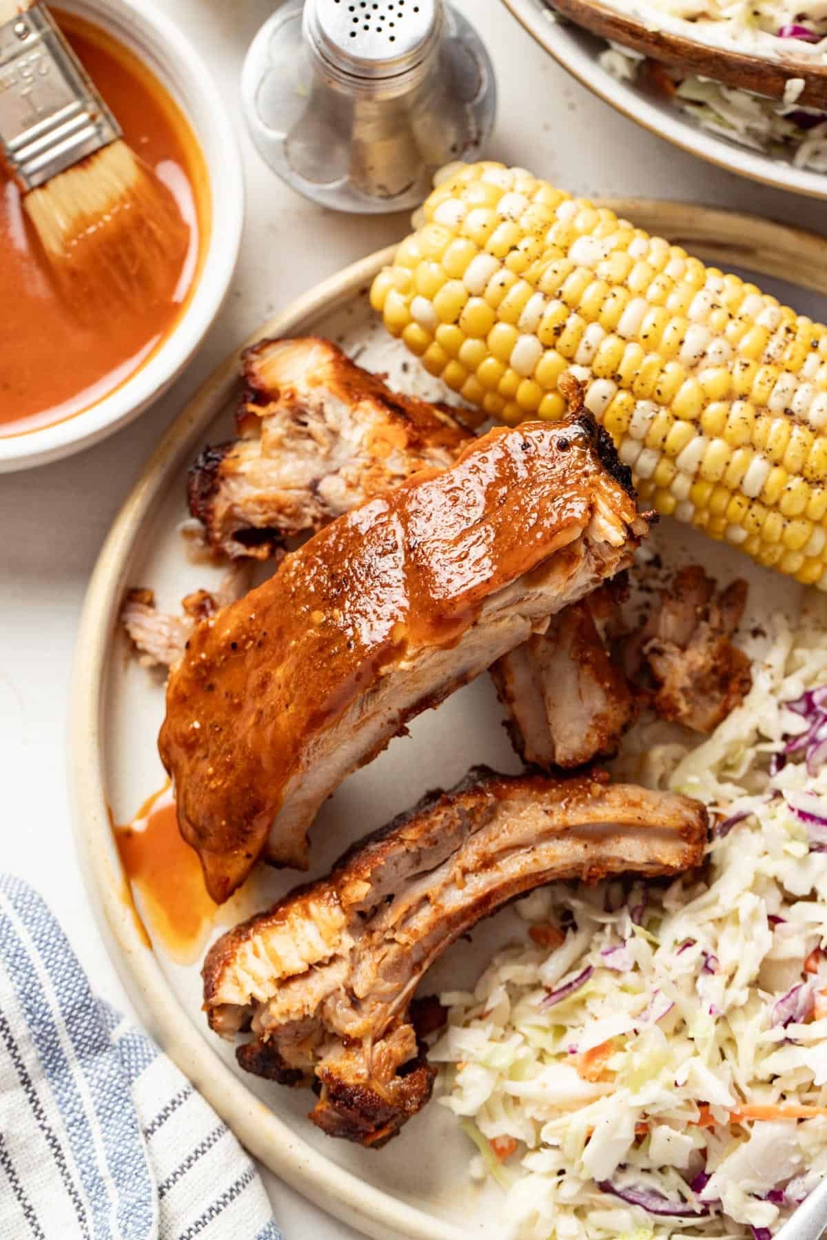 Ribs stacked on a plate with coleslaw and corn on the cob, next to a bowl of homemade bbq sauce.