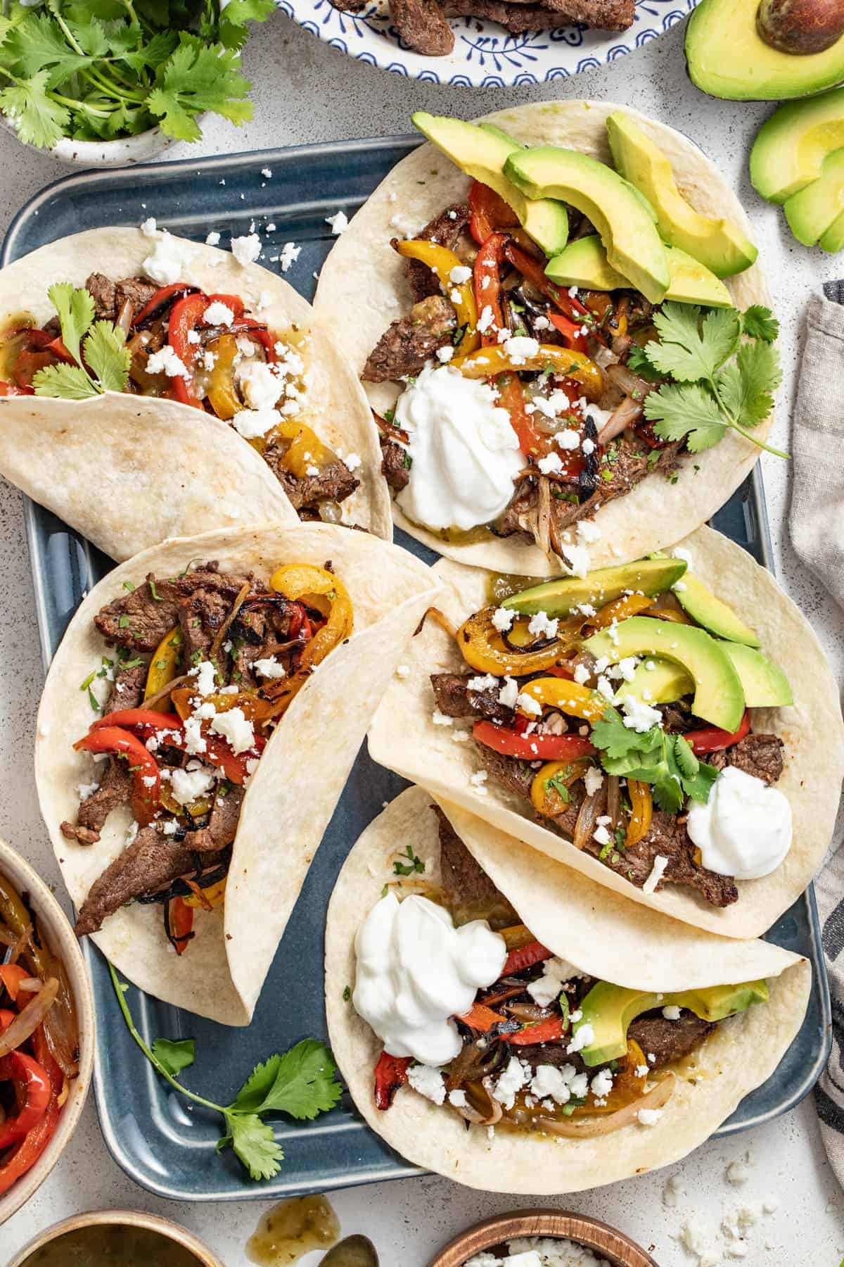 Flank steak fajitas topped with cheese, sour cream and sliced avocados on a gray sheet pan.