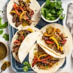 Four fajitas with flank steak, peppers, and shallots on an sheet pan next to a bowl of cilantro.