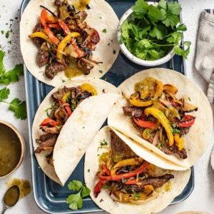 Four fajitas with flank steak, peppers, and shallots on an sheet pan next to a bowl of cilantro.