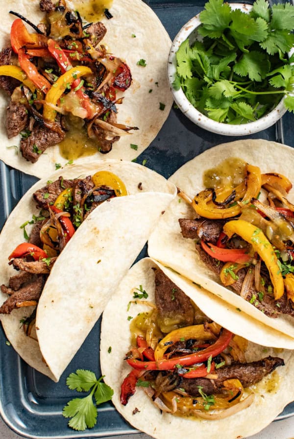 Four fajitas with flank steak, peppers, and shallots on an sheet pan next to a bowl of cilantro.