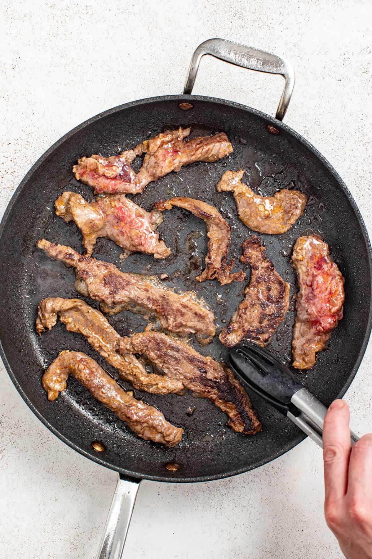 A pan with seared flank steak, and tongs flipping over a strip to show how it should be cooked.