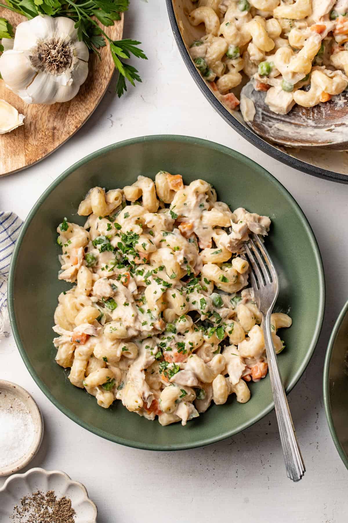 A bowl of pot pie pasta next to a sauce pan with more pasta and fresh garlic and parsley in the corner.