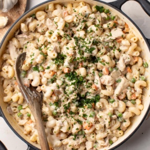 A creamy chicken pot pie pasta in a large cast iron pot with a wood spoon stirring the pasta.