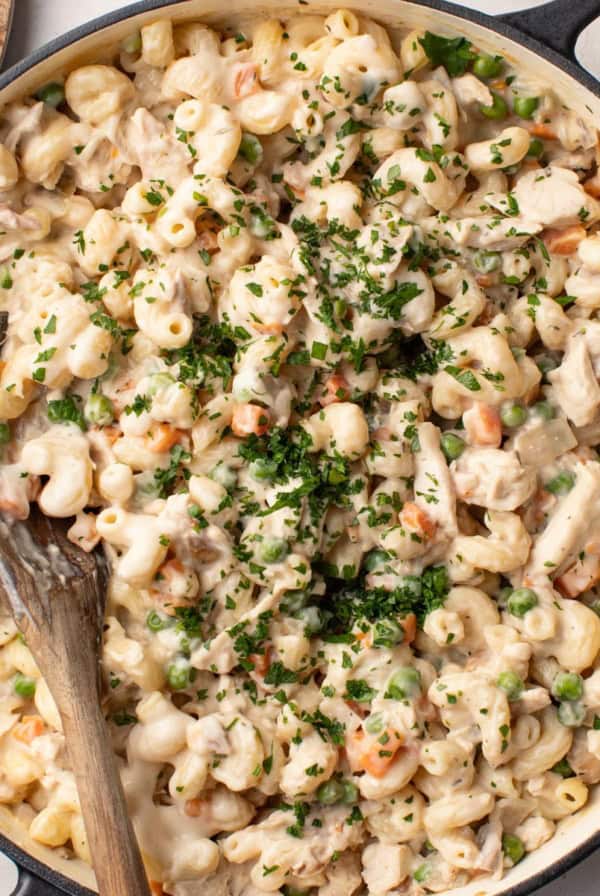 A creamy chicken pot pie pasta in a large cast iron pot with a wood spoon stirring the pasta.