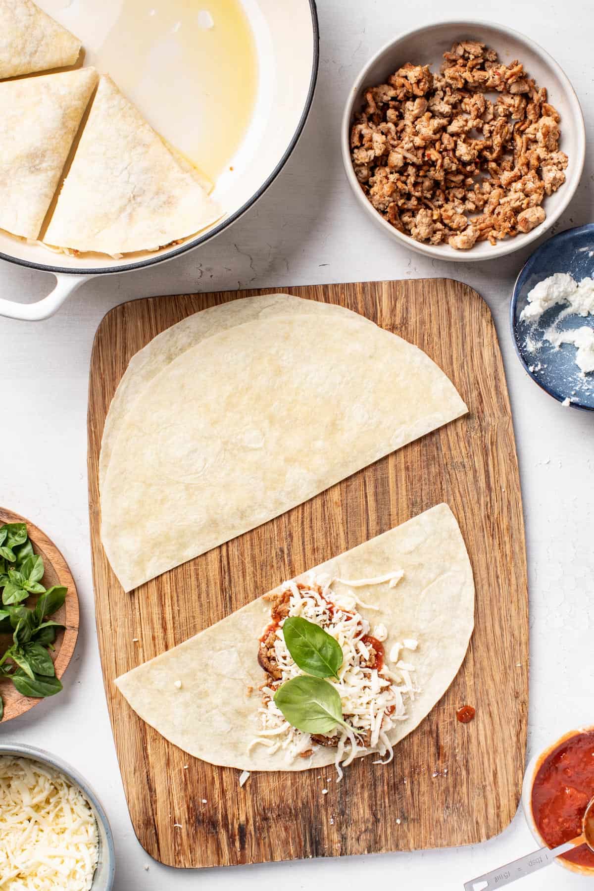 Adding marinara, cheese, and basil to the sliced tortilla in a triangle shape, on the cutting board.