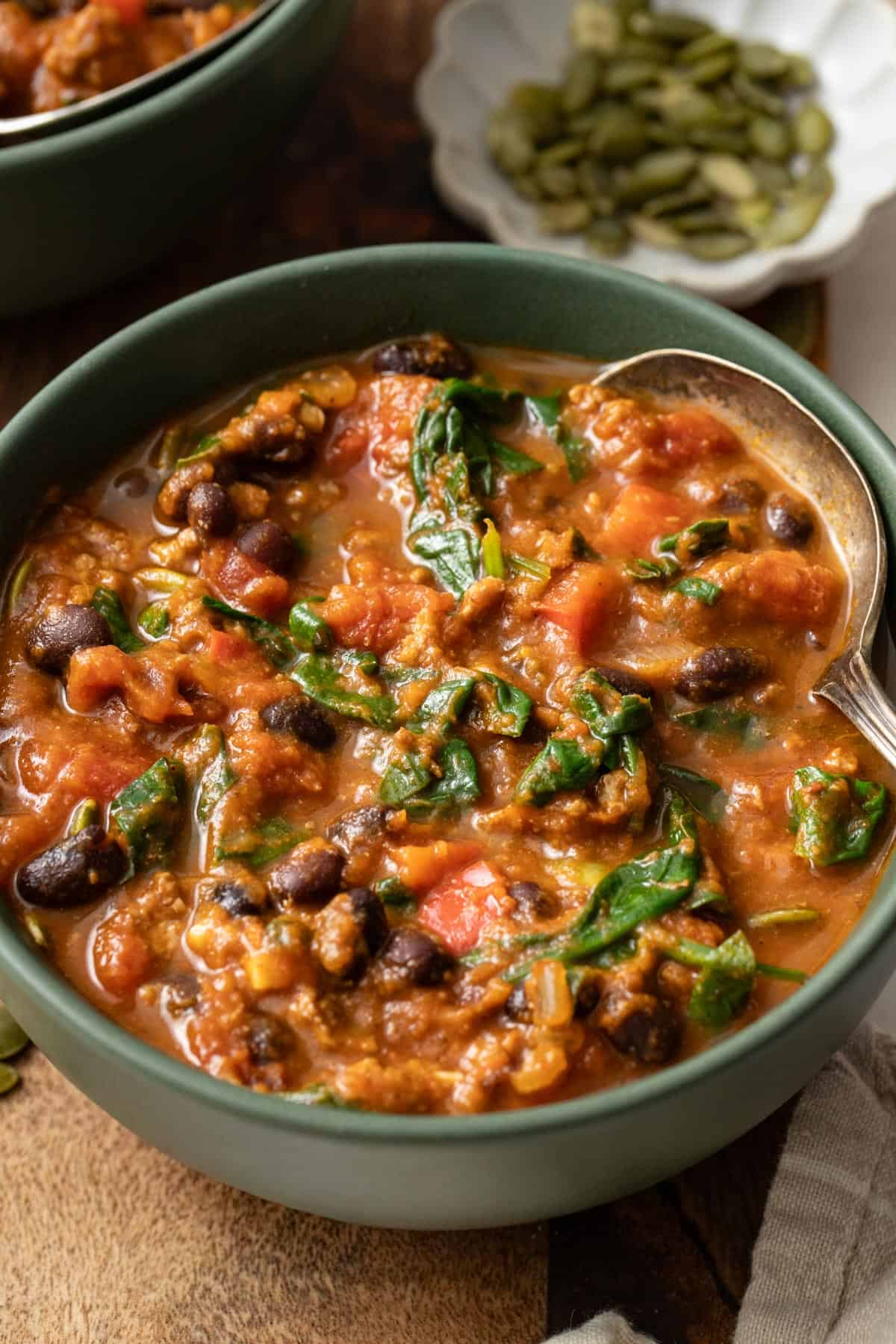 A spoon dipping into a bowl of red chili with black beans, spinach, and tomatoes.