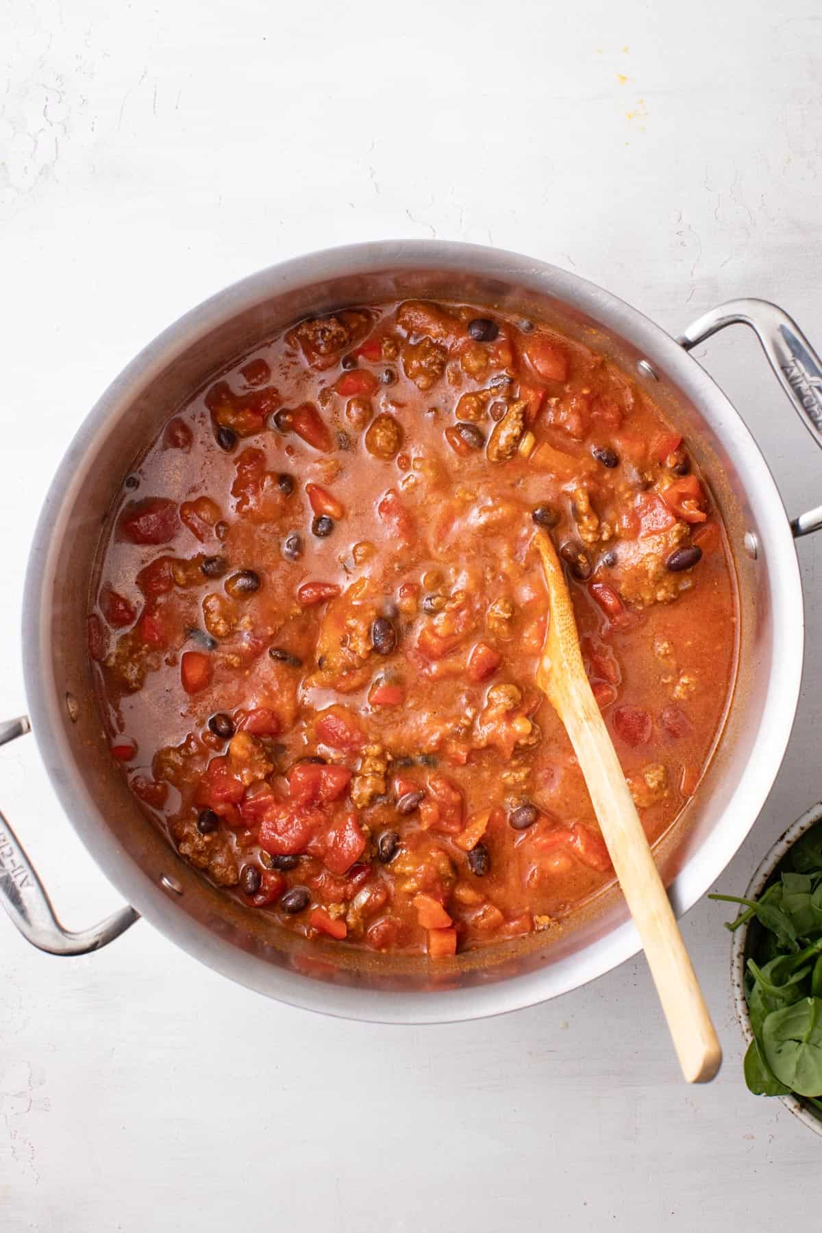 A soup pot with ground beef, chili spices, and broth after being simmered, with a wood spoon dipping into the chili.