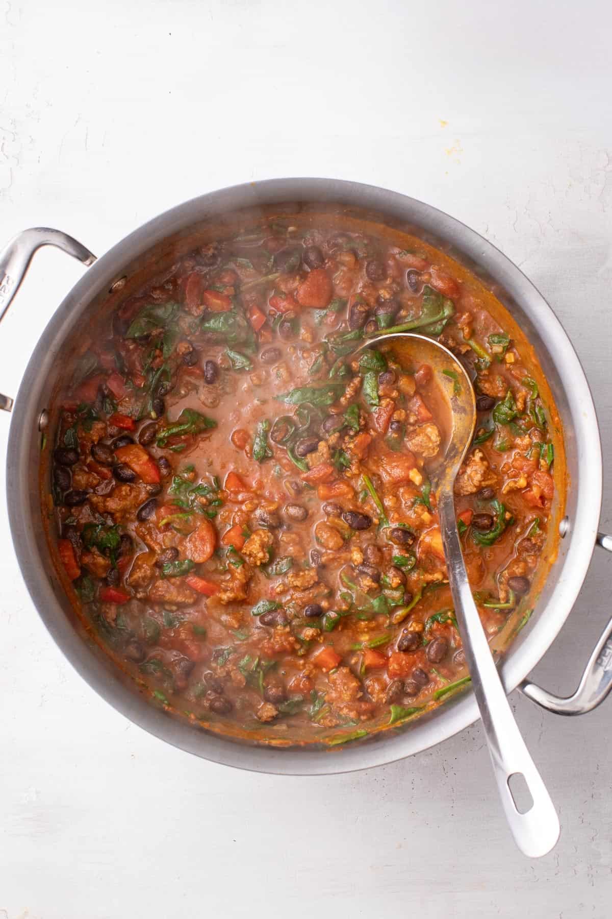 Adding fresh spinach leaves to the chili and stirring with a ladle for serving.