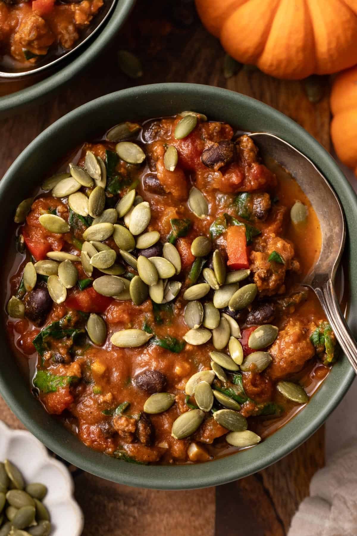 A chili with pumpkin seeds in a green bowl with a spoon dipping in, next to two orange pumpkins.